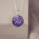 A round sterling silver pendant, hanging from a silver belcher chain, filled with purple crushed crystal and cremation ashes.