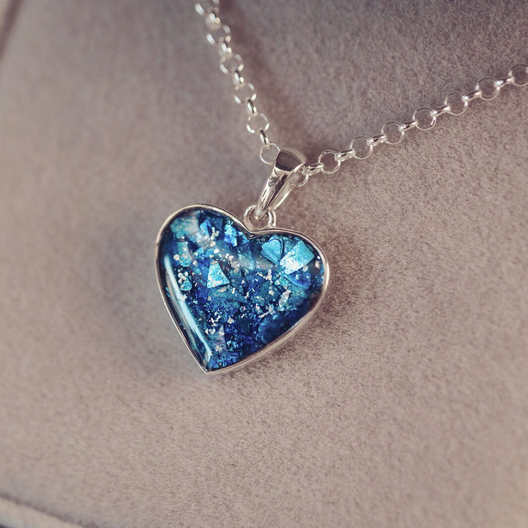 Sterling silver heart necklace with a central section filled with memorial ashes and blue crushed crystals, displayed on a grey fabric background.