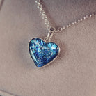 Sterling silver heart necklace with a central section filled with memorial ashes and blue crushed crystals, displayed on a grey fabric background.