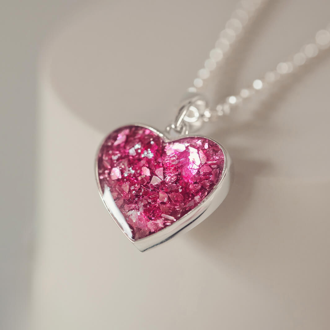 Sterling silver heart necklace with a central section filled with memorial ashes and pink crushed crystals, displayed on a blurred white background