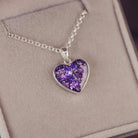 Sterling silver heart necklace with a central section filled with memorial ashes and purple crushed crystals, displayed on a grey fabric background.