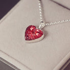 Sterling silver heart necklace with a central section filled with memorial ashes and red crushed crystals, displayed on a grey fabric background.