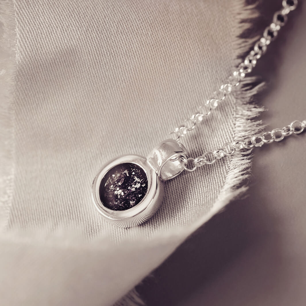 Sterling silver round necklace with a central section filled with memorial ashes and black crushed crystals, displayed on a grey fabric