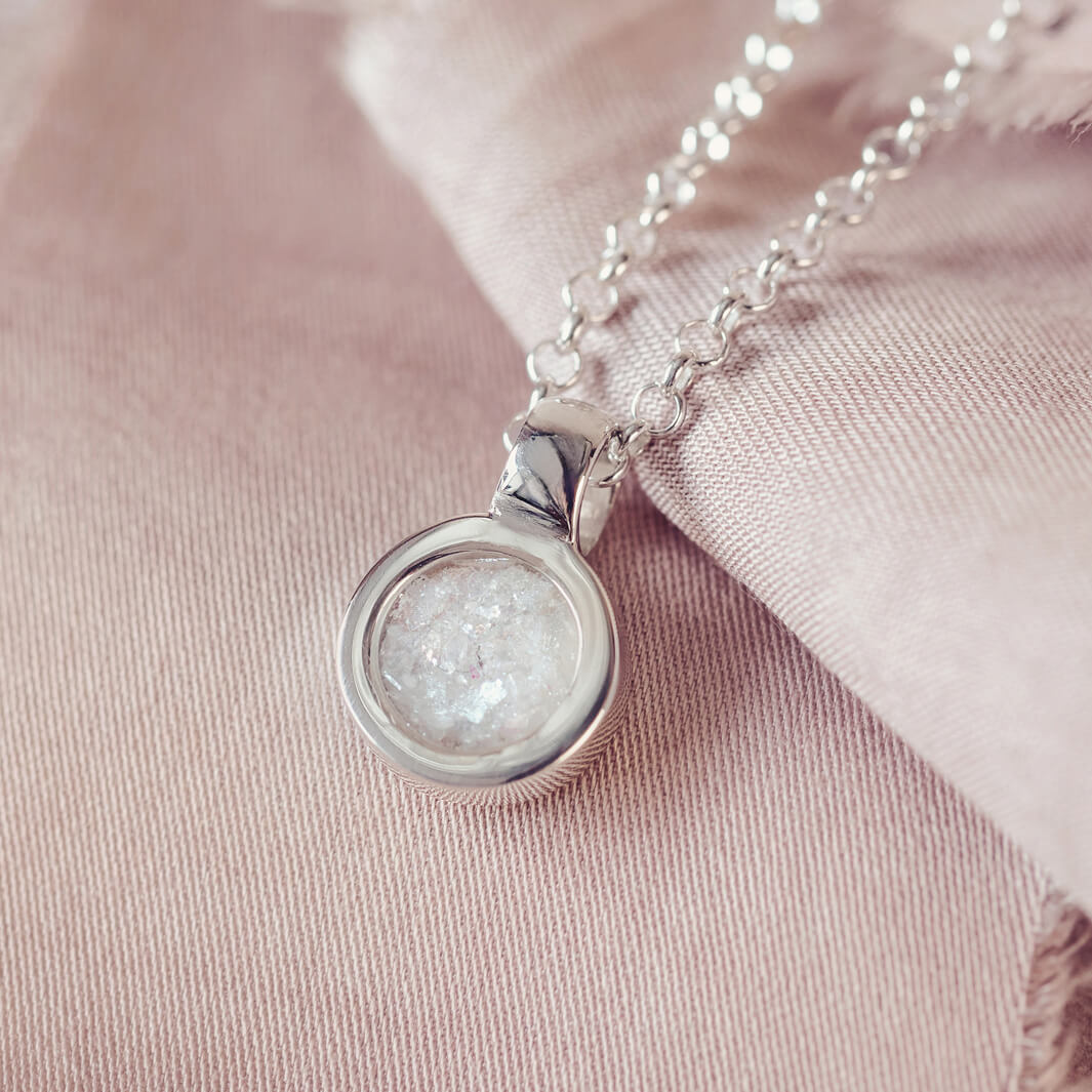 Sterling silver round necklace with a central section filled with memorial ashes and clear crushed crystals, displayed on a pink background