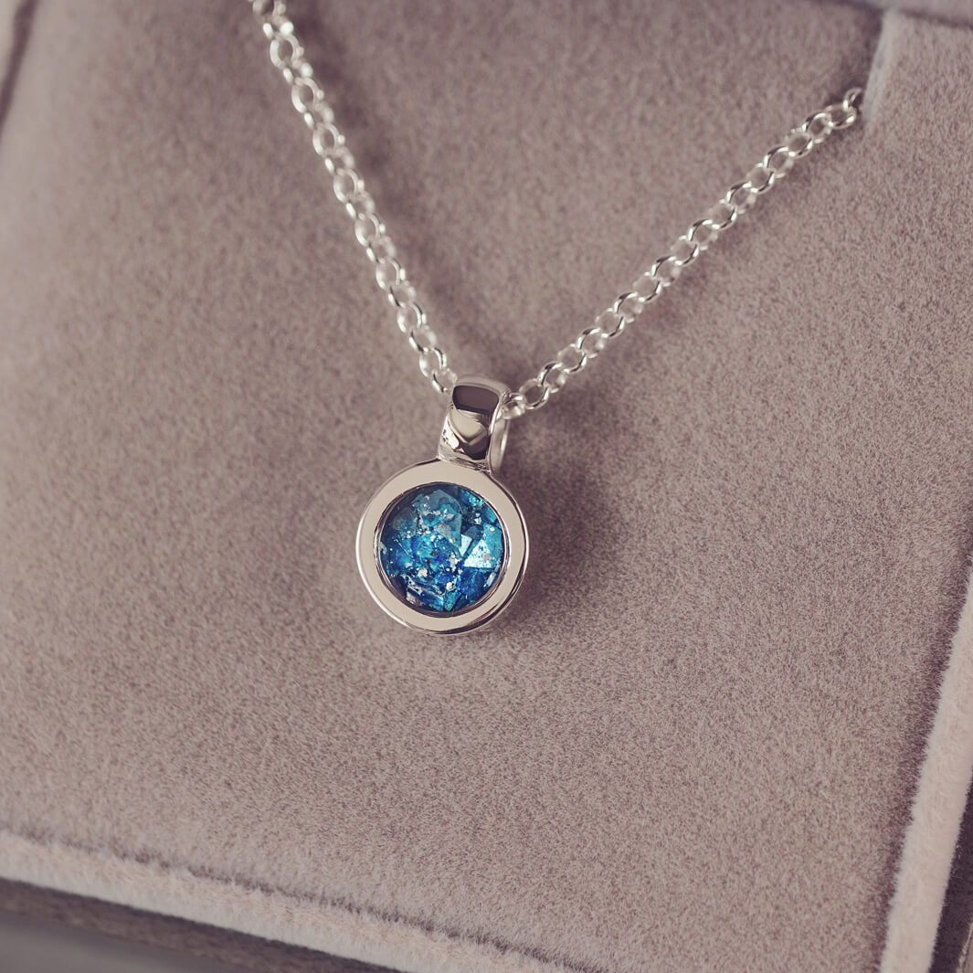 Sterling silver small round necklace with a central section filled with blue memorial ashes and crushed crystals, displayed on a beige fabric background.