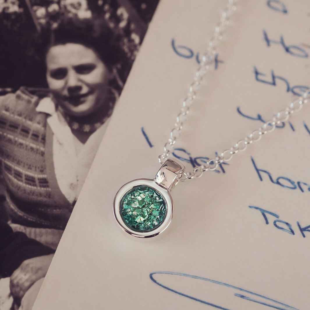 Sterling silver round necklace with a central section filled with memorial ashes and green crushed crystals, displayed on a background of a handwritten message and photograph