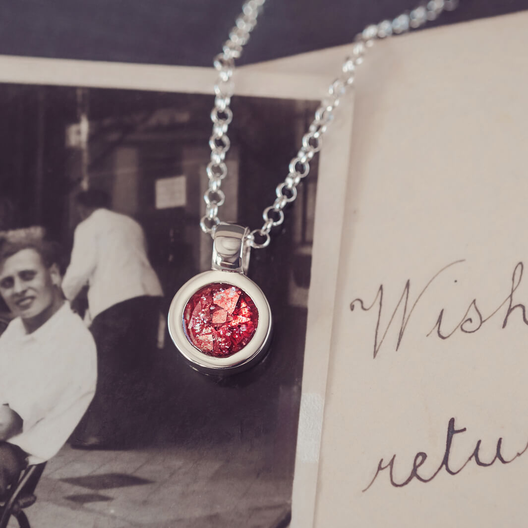 Sterling silver round necklace with a central section filled with memorial ashes and red crushed crystals, displayed on a background of a handwritten message and photograph