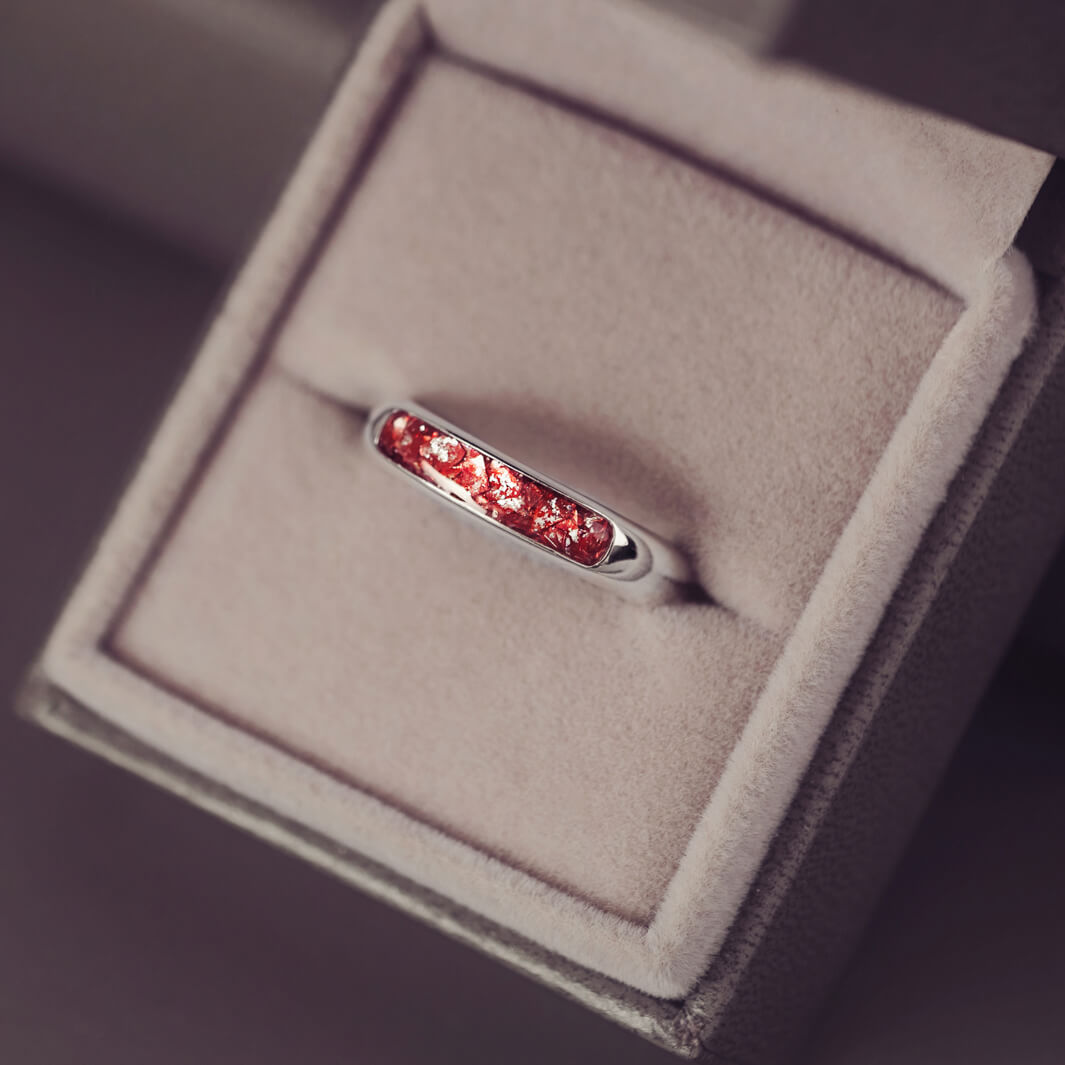 Sterling silver womens ring band with a central section filled with  memorial ashes and red crushed crystals, displayed in a grey jewellery box