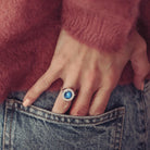 The image shows a close-up of a person’s hand resting near their back pocket, with their fingers partially tucked into blue denim jeans. They are wearing a large, silver ring featuring a vibrant blue stone containing cremation ashes and resin, surrounded by a halo of smaller stones. The person is dressed in a soft, textured, rose-coloured sweater, and the rich colours of the sweater and the blue stone create a striking contrast. The ring stands out as a bold and elegant piece.