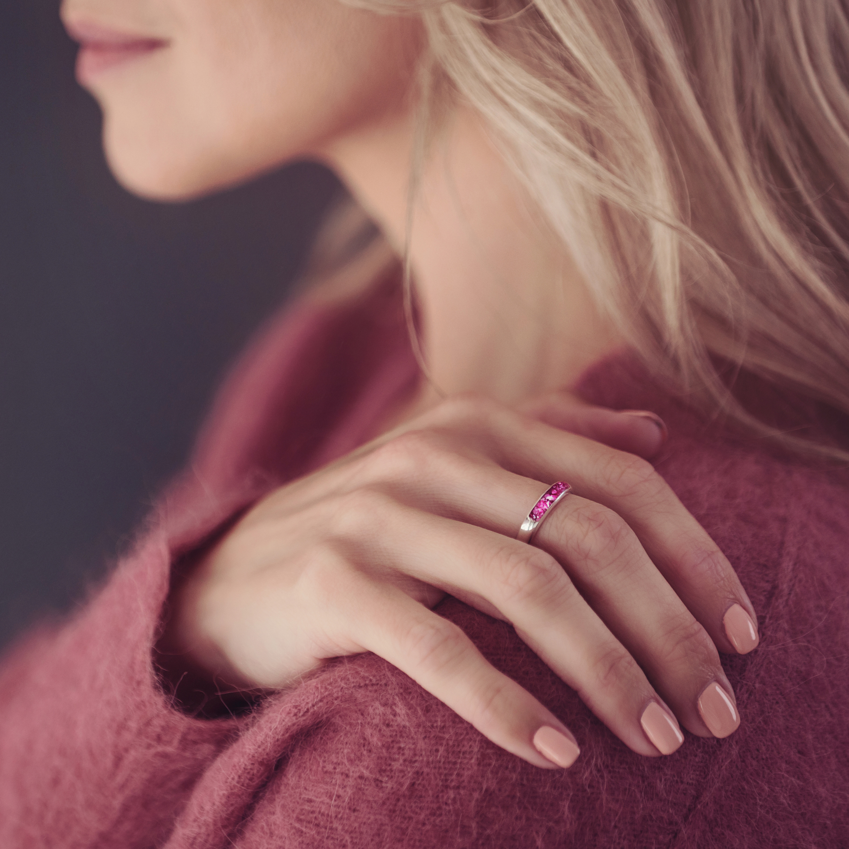 The image shows a close-up of a person’s hand resting on their shoulder. They are wearing a silver ring with a band of bright pink stone, containing cremation ashes and resin. The person is dressed in a soft, textured, rose-coloured sweater that complements the colour of the stone. Their nails are neatly manicured with a neutral polish. The soft lighting highlights the ring, drawing attention to its vibrant colour and elegant design, suggesting that it holds sentimental or memorial significance. 