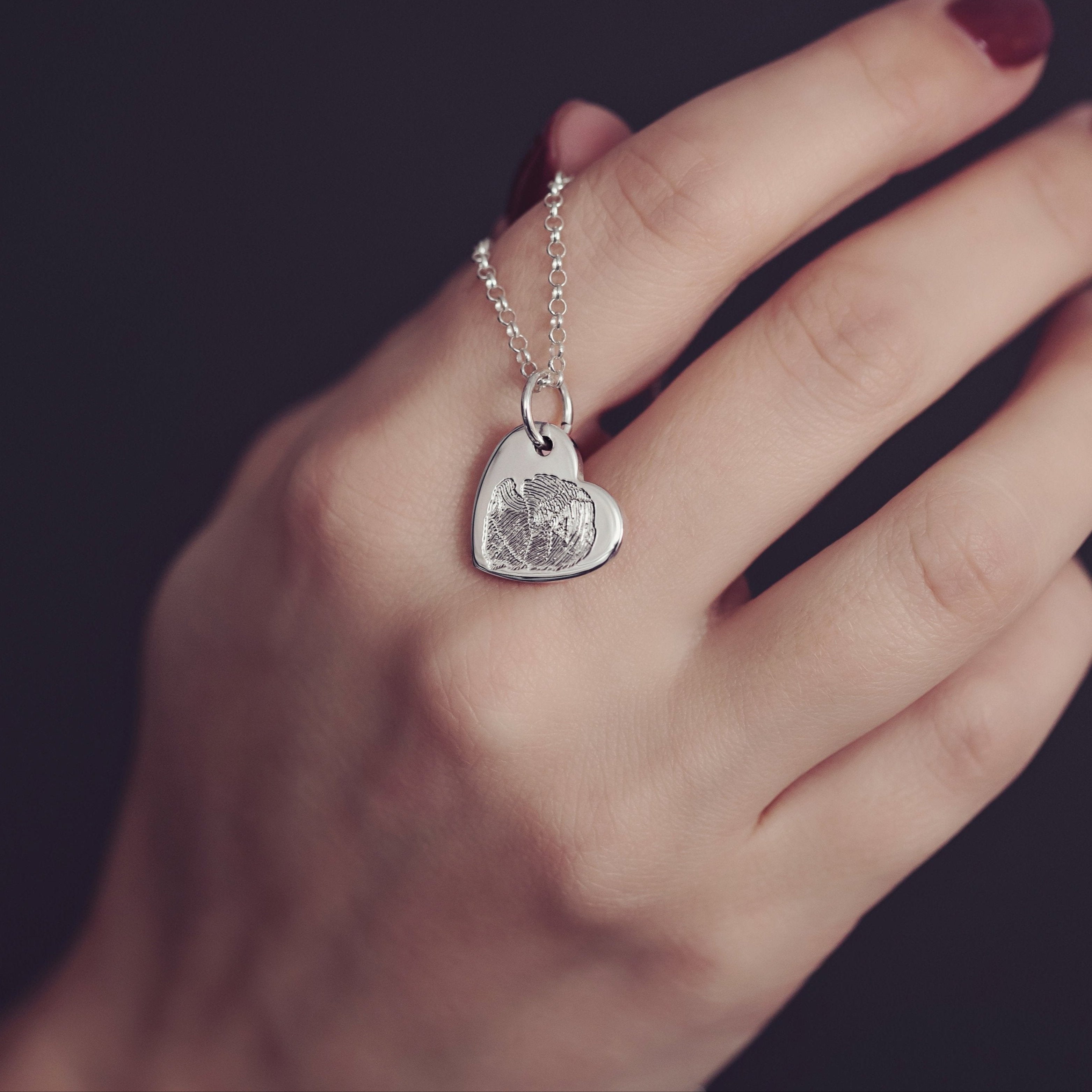 A woman's hand delicately holds a silver heart necklace featuring a unique fingerprint detail