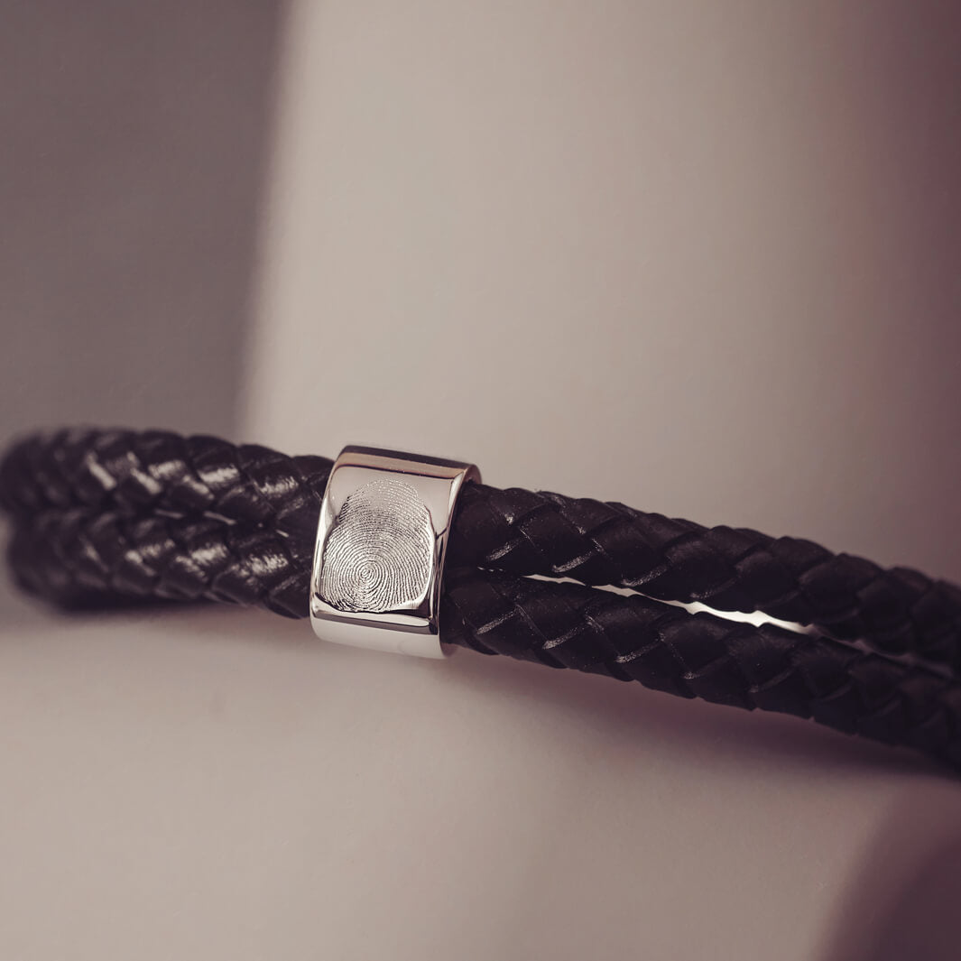 Black braided leather bracelet featuring a silver charm engraved with a fingerprint, displayed in a grey jewellery box