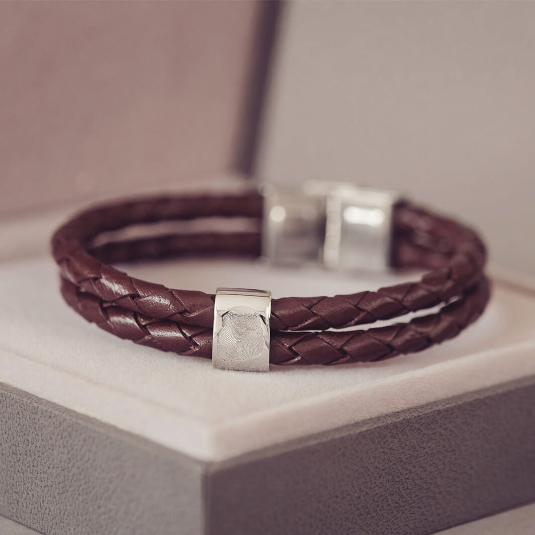 Brown braided leather bracelet featuring a silver charm engraved with a fingerprint, displayed in a grey jewellery box