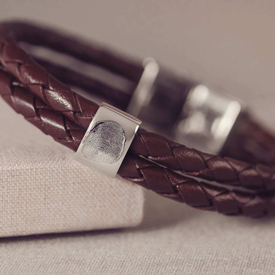 Brown braided leather bracelet featuring a silver charm engraved with a fingerprint, displayed in a grey jewellery box