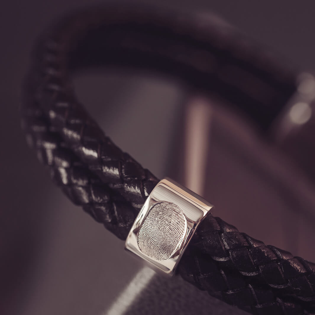 Black braided leather bracelet featuring a silver charm engraved with a fingerprint, displayed in a grey jewellery box