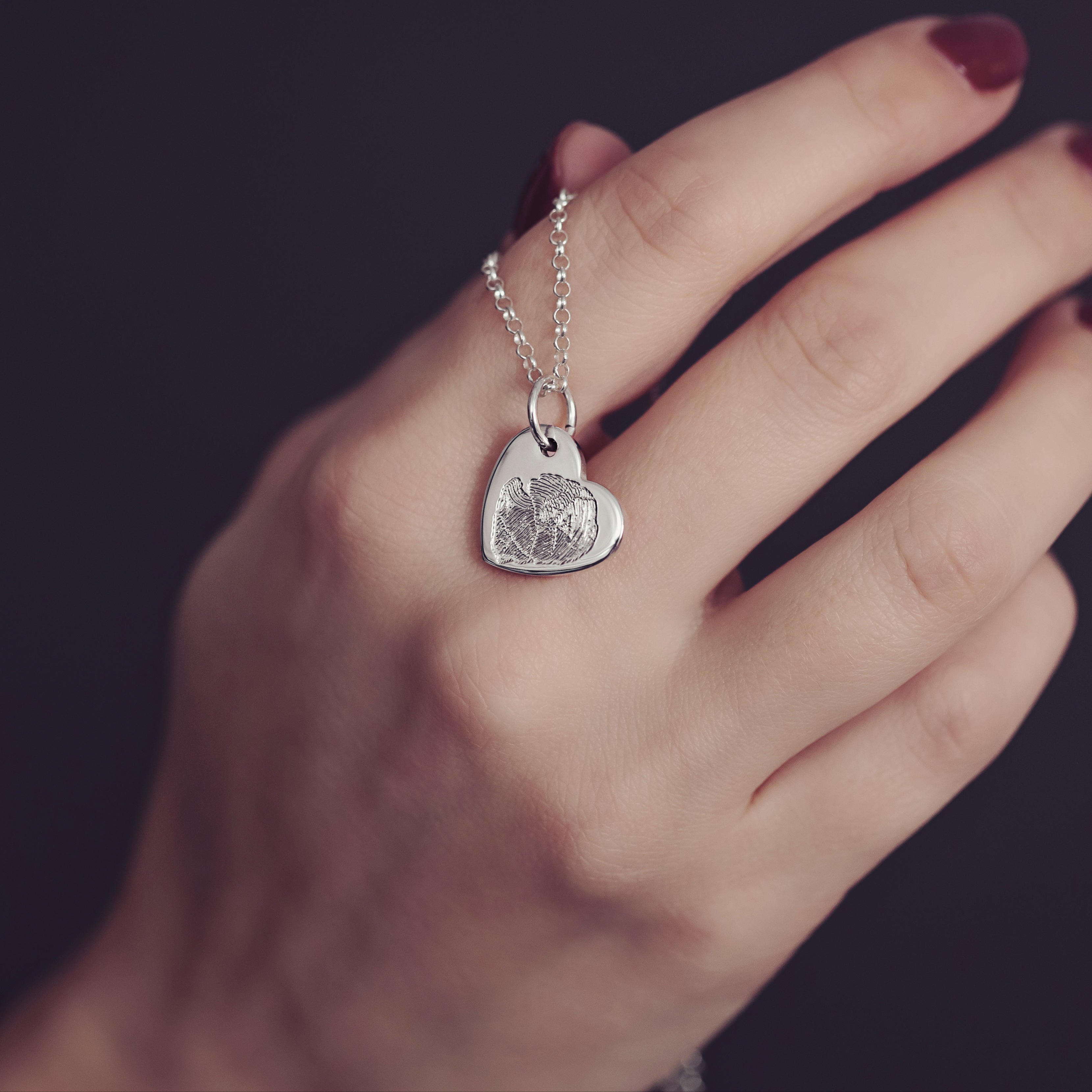 A woman's hand delicately holds a sterling silver heart necklace featuring a fingerprint