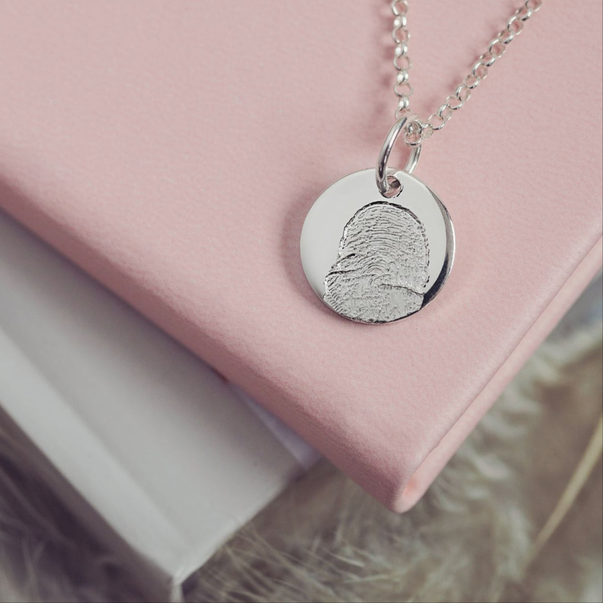 Sterling silver round fingerprint necklace resting on a soft pink leather book