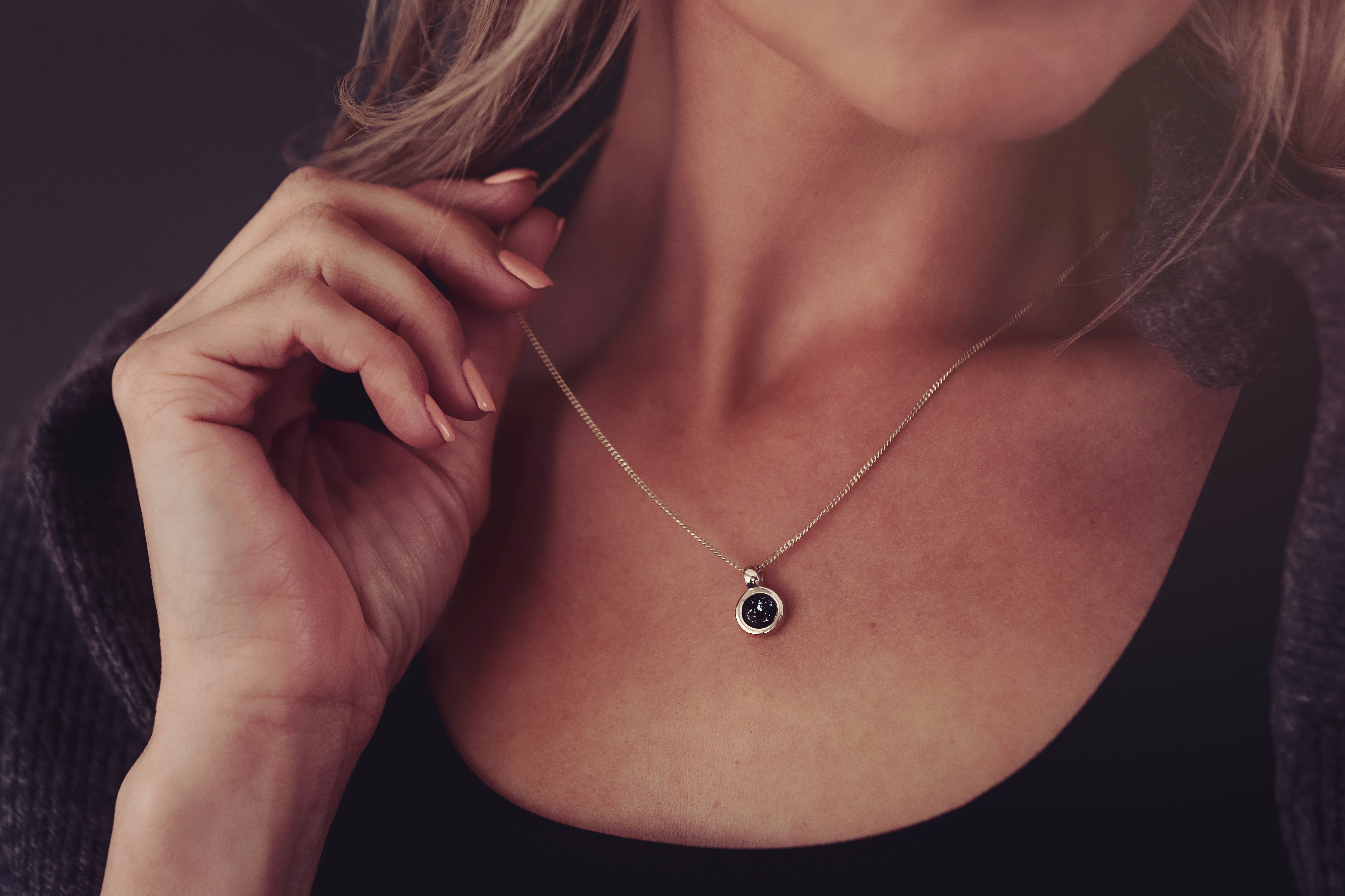 Close up of a woman wearing a delicate gold round necklace, featuring a small black pendant containing cremation ashes