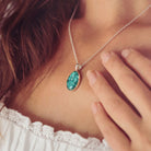 Neckline of a lady with brown hair wearing a Cremation Ashes Oval Necklace, filled with ashes and green crushed crystal. The lady is wearing a white top and is holding her hand towards the necklace