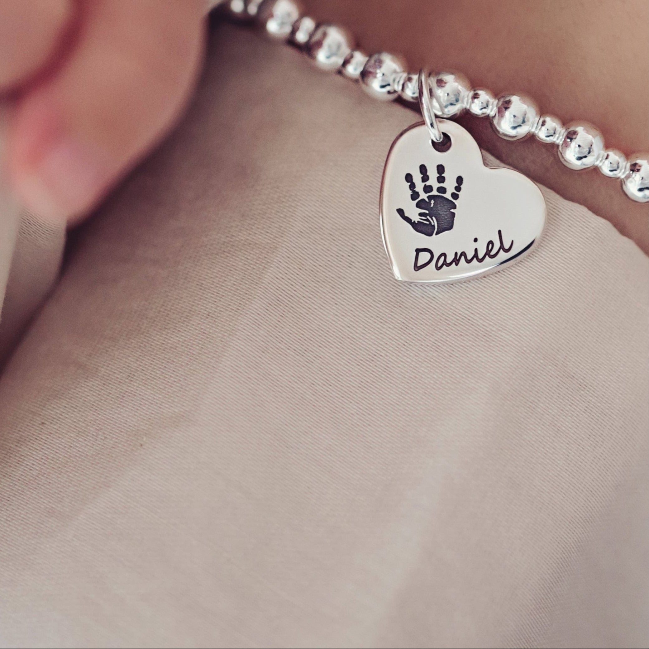 A close-up of a woman holding a baby's hand, featuring a handprint bracelet with the name 'Daniel' engraved