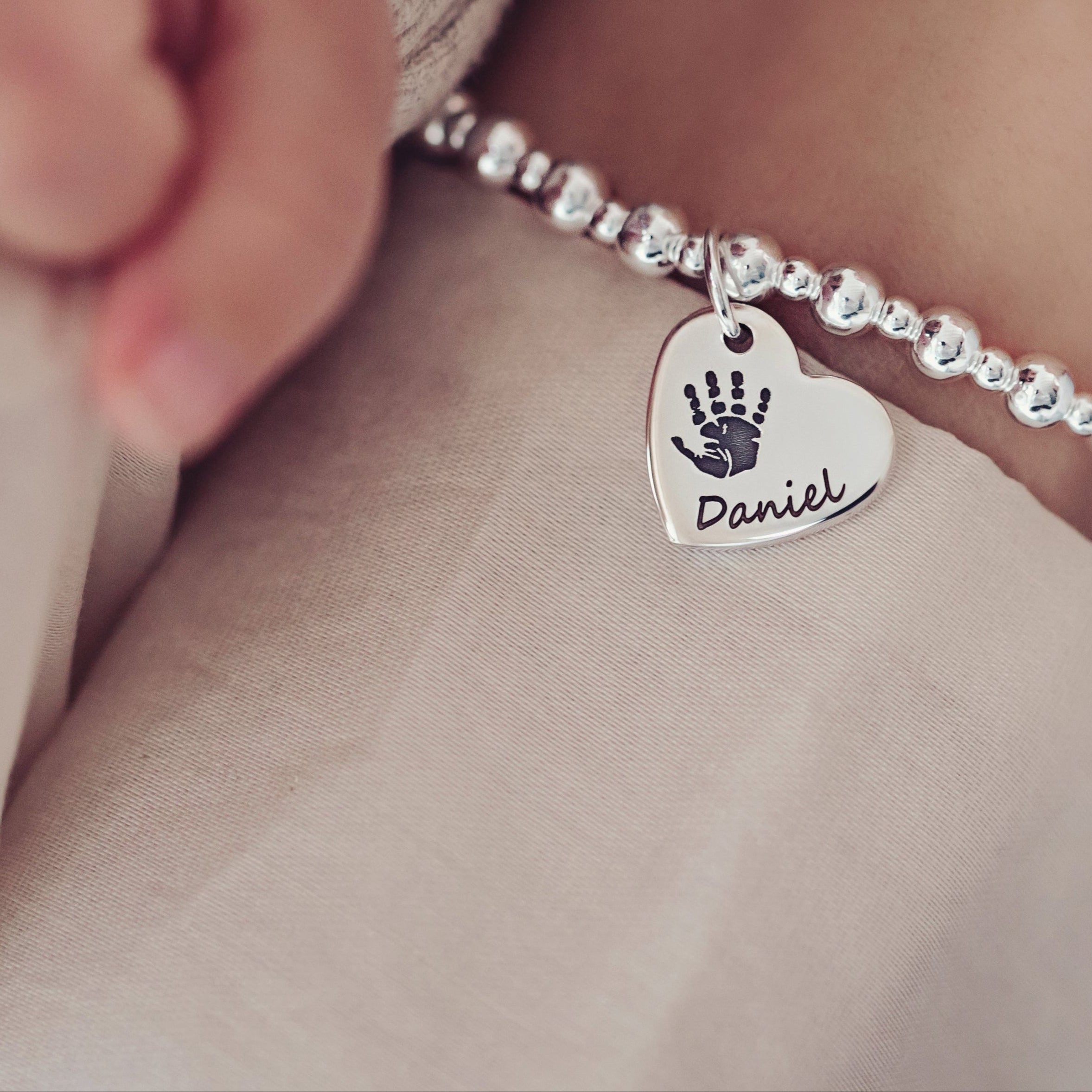 A close-up of a woman holding a baby's hand, featuring a handprint bracelet with the name 'Daniel' engraved