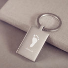 A stainless steel rectangular keyring, engraved with one footprint and name, leaning on a fabric background