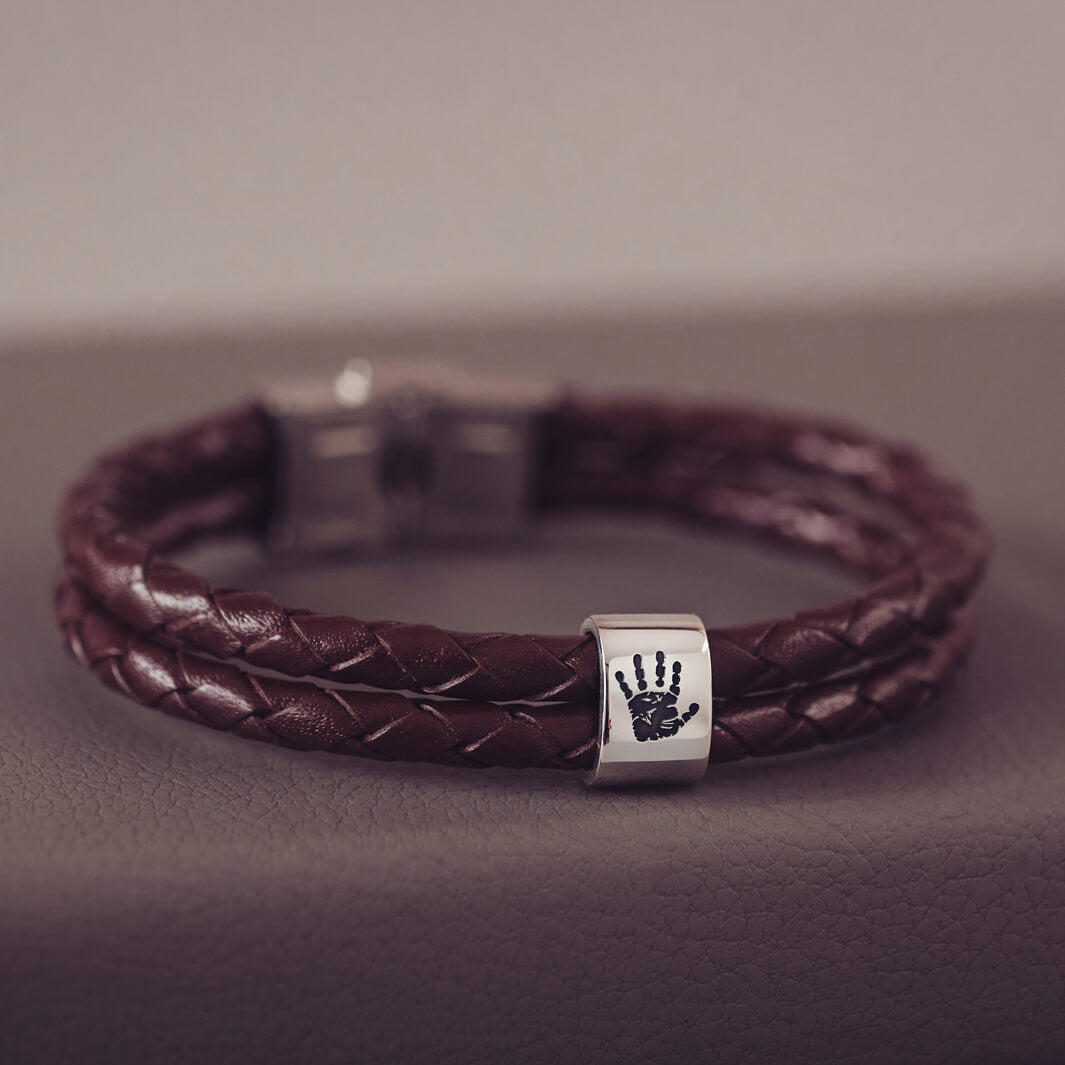 Brown braided leather bracelet featuring a sterling silver charm engraved with a black handprint, displayed on a soft grey background.