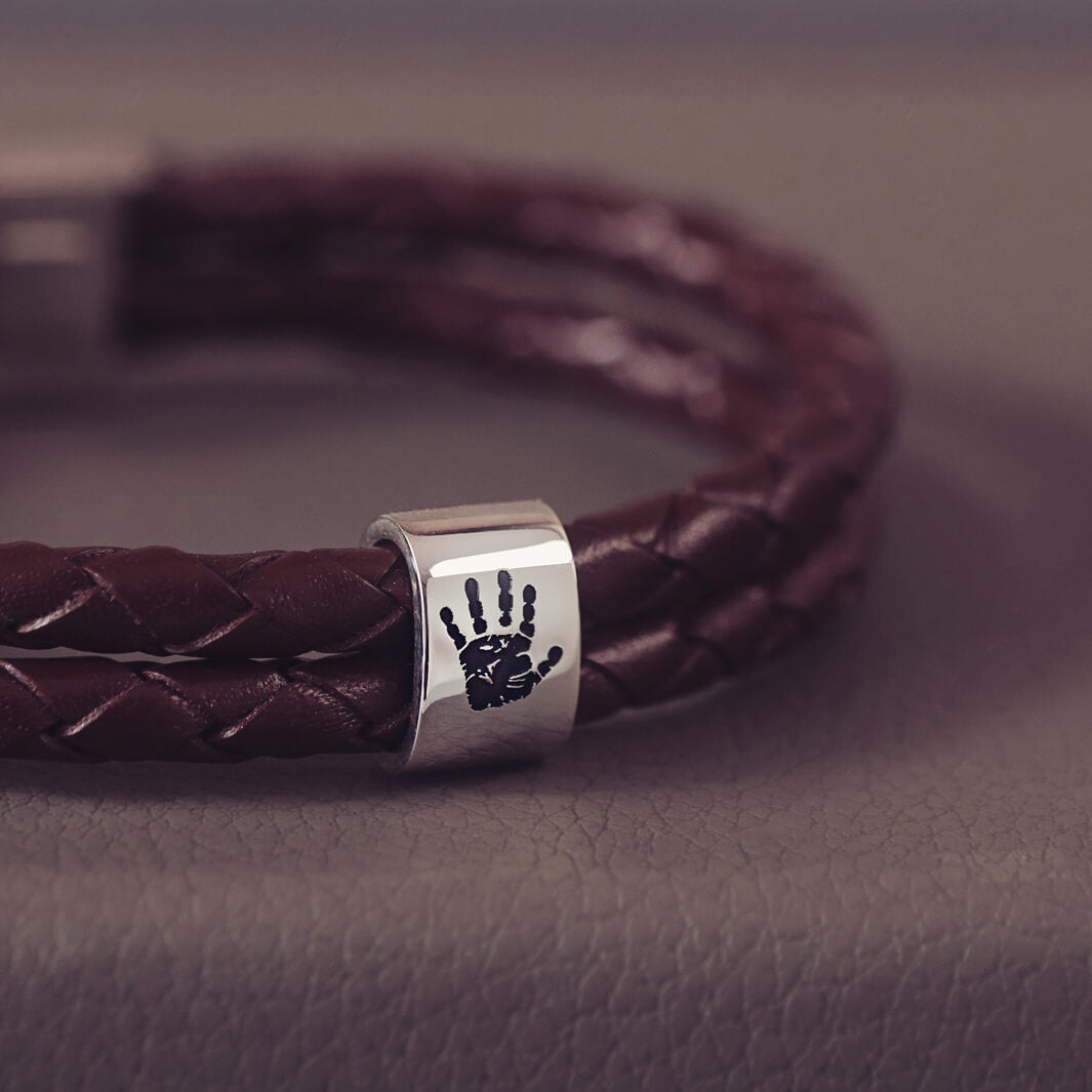 Brown braided leather bracelet featuring a sterling silver charm engraved with a black handprint, displayed on a soft grey background.