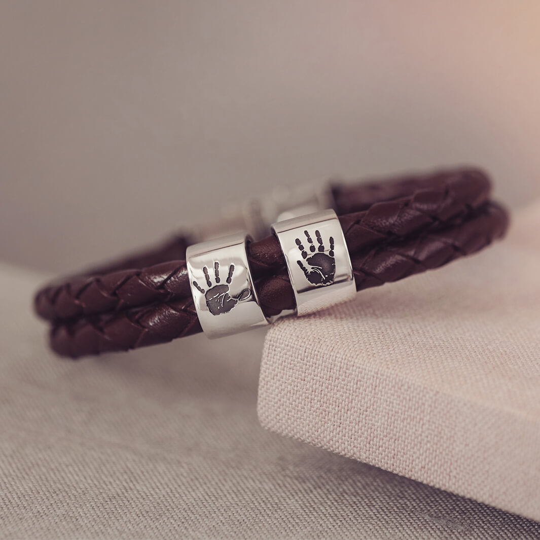 Brown braided leather bracelet featuring two sterling silver charms, each engraved with a black handprint, displayed on a soft grey background.