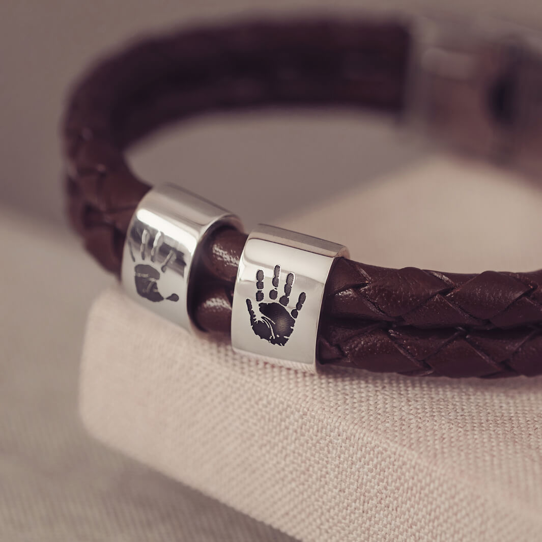 Brown braided leather bracelet featuring two sterling silver charms, each engraved with a black handprint, displayed on a soft grey background.