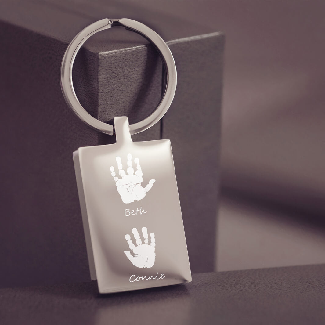 A stainless steel rectangular keyring resting on a dark grey jewellery box, with two handprints and names engraved onto the keyring