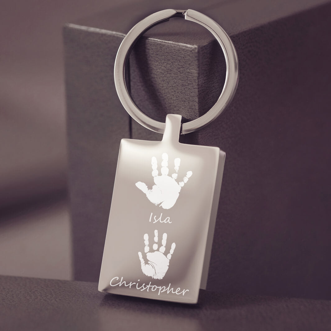 A stainless steel rectangular keyring resting on a dark grey jewellery box, with two handprints and names engraved onto the keyring