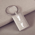 A stainless steel rectangular keyring, engraved with two footprints and names, leaning on a fabric background