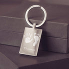 A stainless steel keyring, engraved with a handprint, footprint and name, leaning against a grey jewellery box