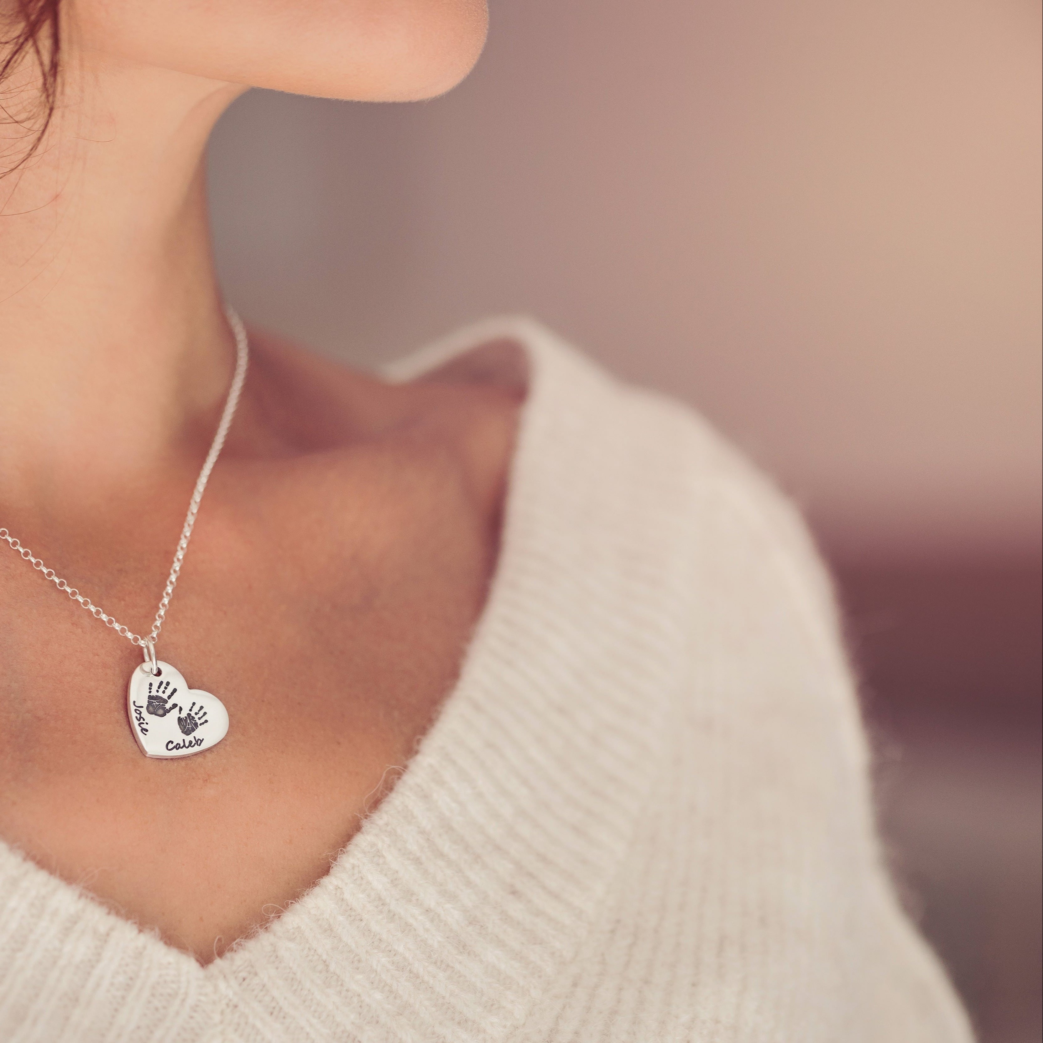 A woman in a white jumper wearing a silver heart necklace featuring two handprints and names 'Josie' and 'Caleb'