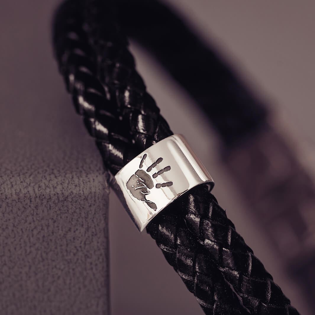 Black braided leather bracelet featuring a sterling silver charm engraved with a black handprint, displayed on a soft grey background.