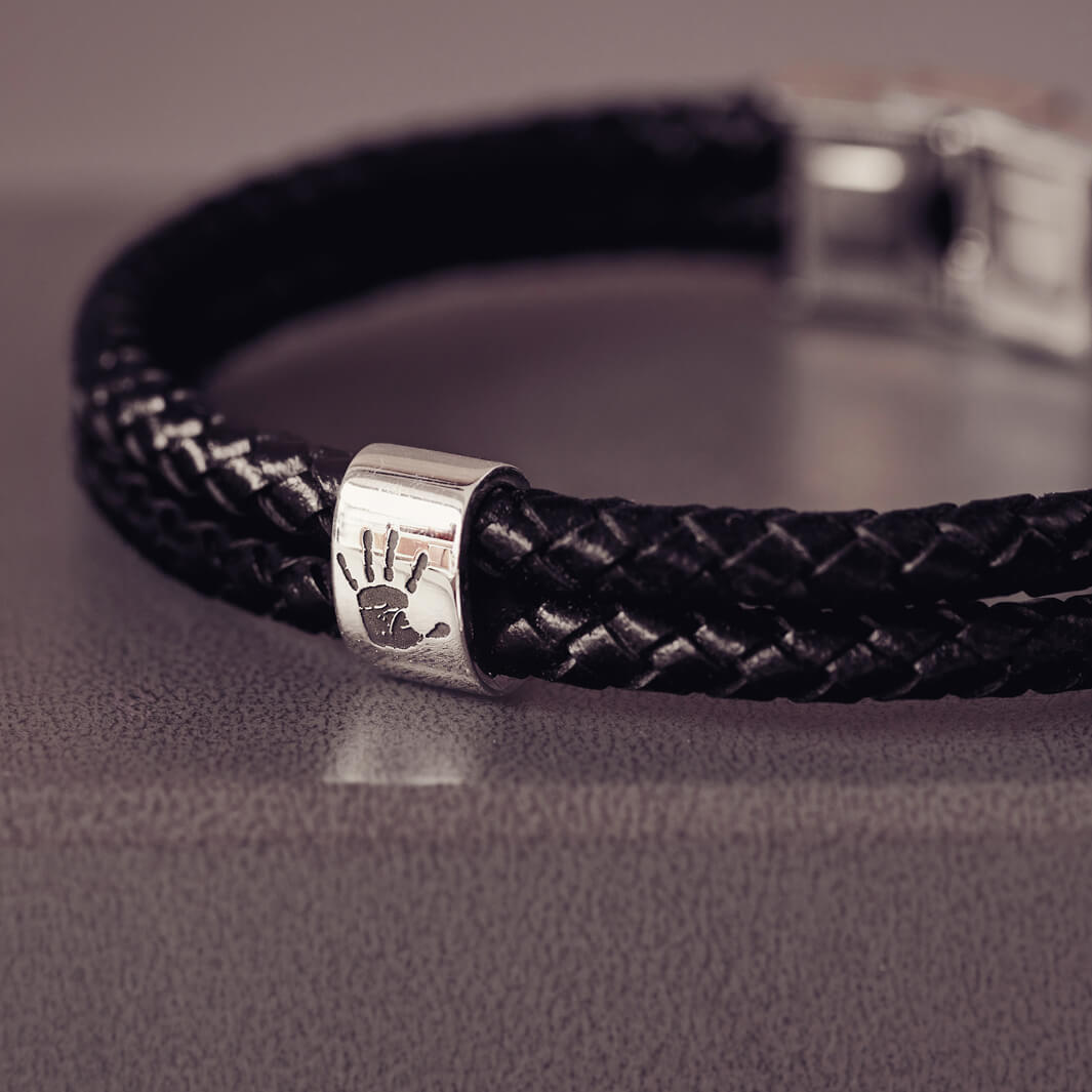 Black braided leather bracelet featuring a sterling silver charm engraved with a black handprint, displayed on a soft grey background.