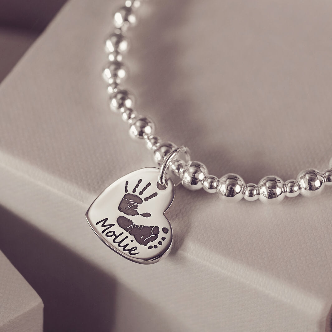 Silver beaded bracelet featuring a heart-shaped charm engraved with a handprint and footprint, along with the name 'Mollie,' displayed on a grey surface