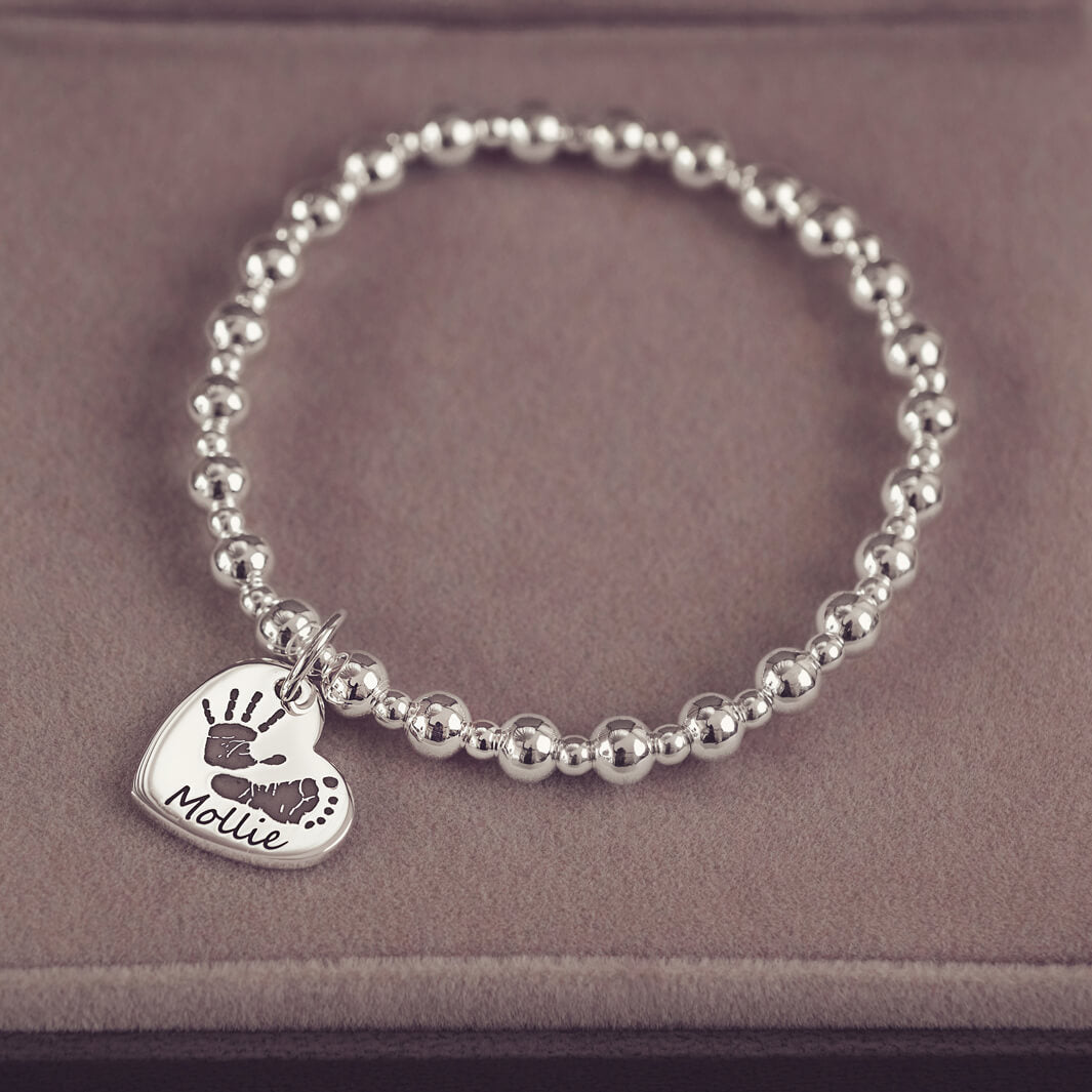 Silver beaded bracelet featuring a heart-shaped charm engraved with a handprint and footprint, along with the name 'Mollie,' displayed on a soft beige surface