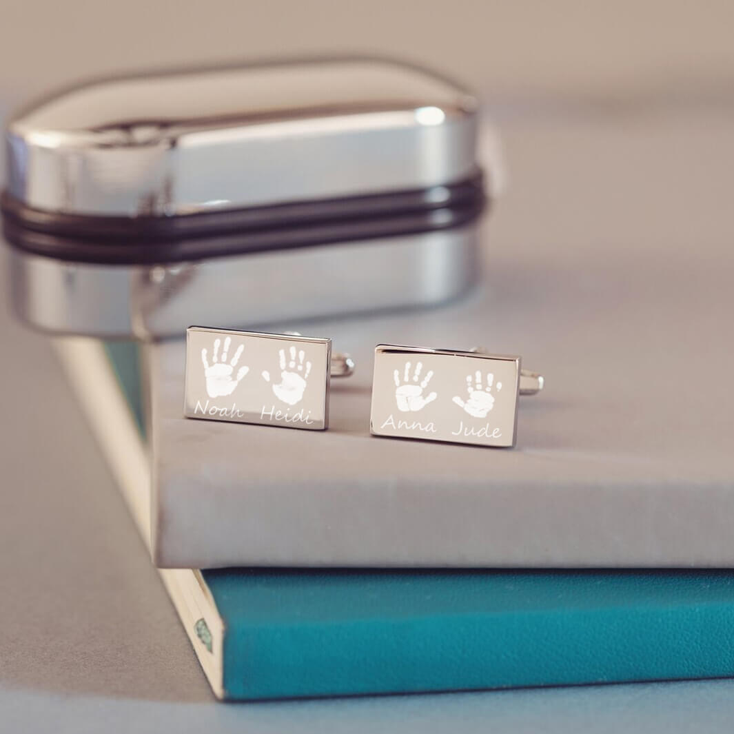 stainless steel rectangular cufflinks with two handprints and names engraved onto each cufflink. Displayed on a grey and blue background, with a chrome cufflink case in the background