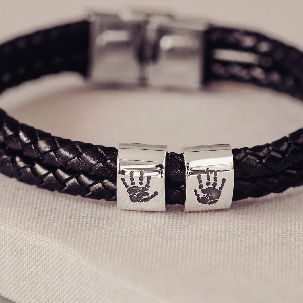 Black braided leather bracelet featuring two sterling silver charms, each engraved with a black handprint, displayed on a soft grey background.