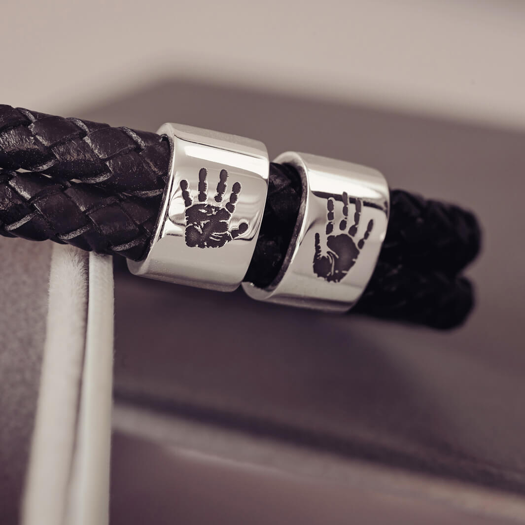 Black braided leather bracelet featuring two sterling silver charms, each engraved with a black handprint, displayed on a soft grey background.