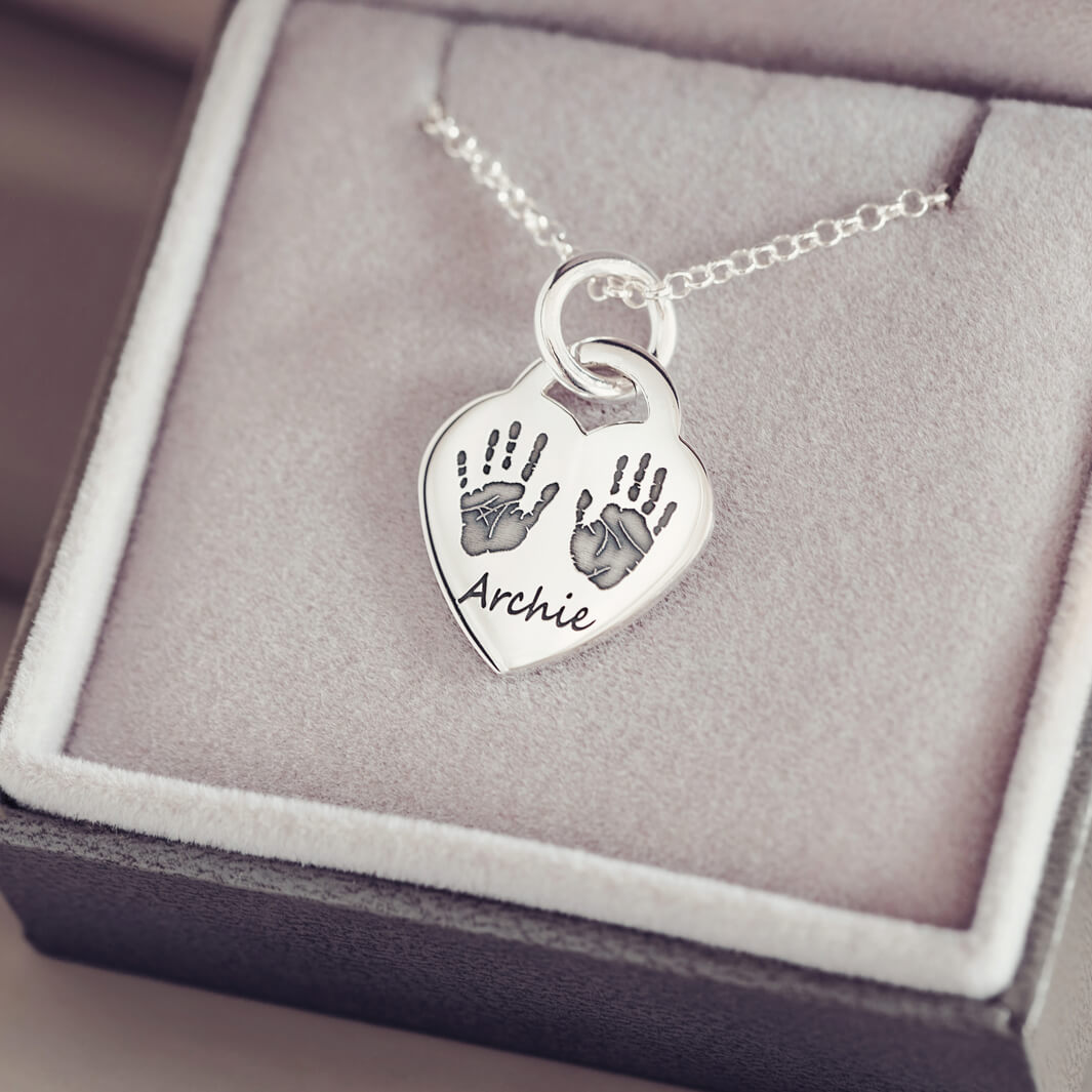 A silver lock heart necklace, on a silver chain, with two black handprint and the name 'Archie' impressed into the front of the necklace. Displayed in a grey jewellery box