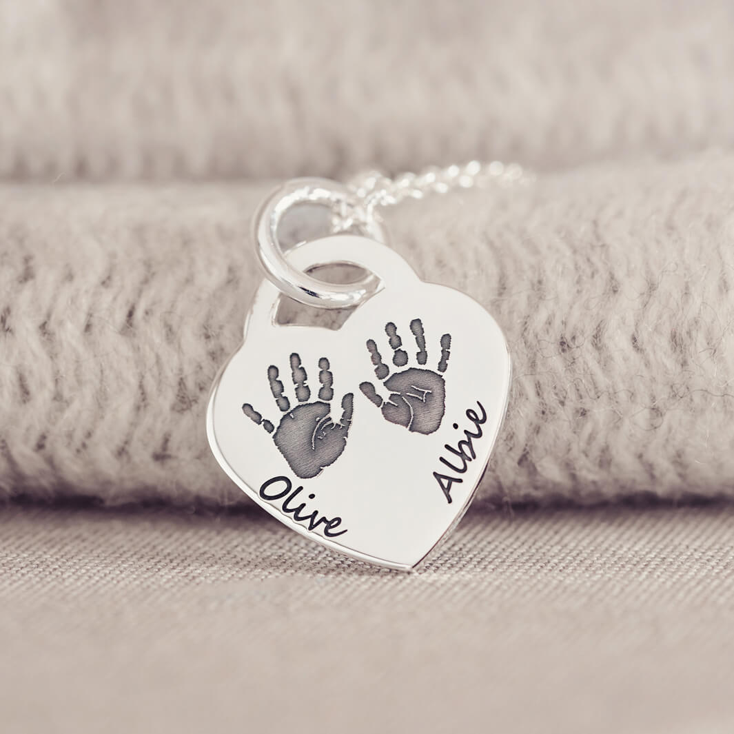 A silver heart shaped necklace, on a silver chain, with two handprints and names Olive and Albie engraved onto the front of the pendant. Displayed on a beige fabric background