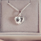 A silver heart shaped hinged locket, on a silver belcher chain, with a dark grey hand and footprint engraved into the front of the locket pendant. Displayed in a grey jewellery box