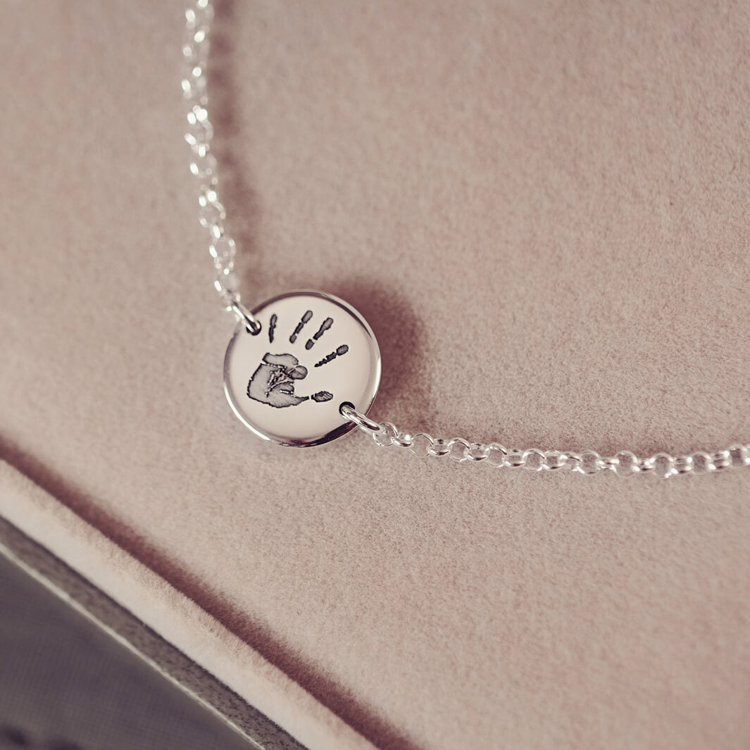 A round silver pendant bracelet featuring a detailed engraved handprint, displayed on a delicate silver chain. The necklace rests in an elegant, soft velvet jewellery box