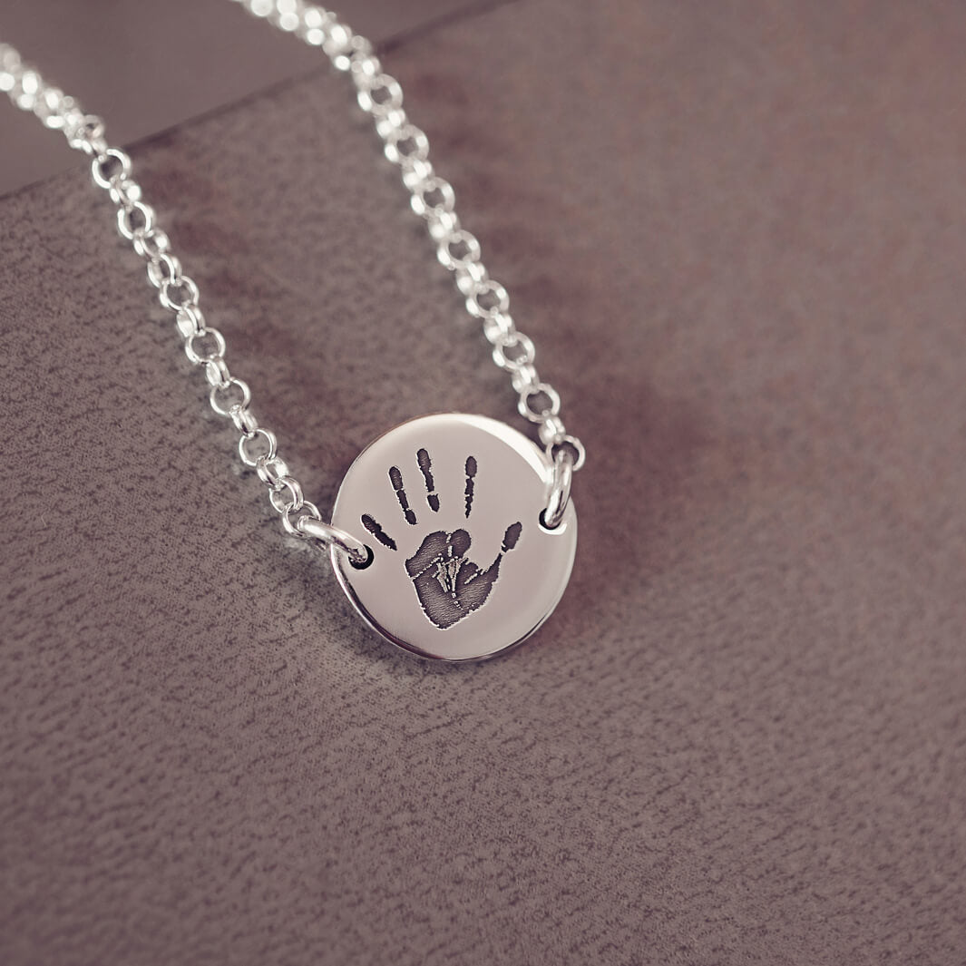 A round silver pendant bracelet featuring a detailed engraved handprint, displayed on a delicate silver chain. The necklace rests in an elegant, soft velvet jewellery box