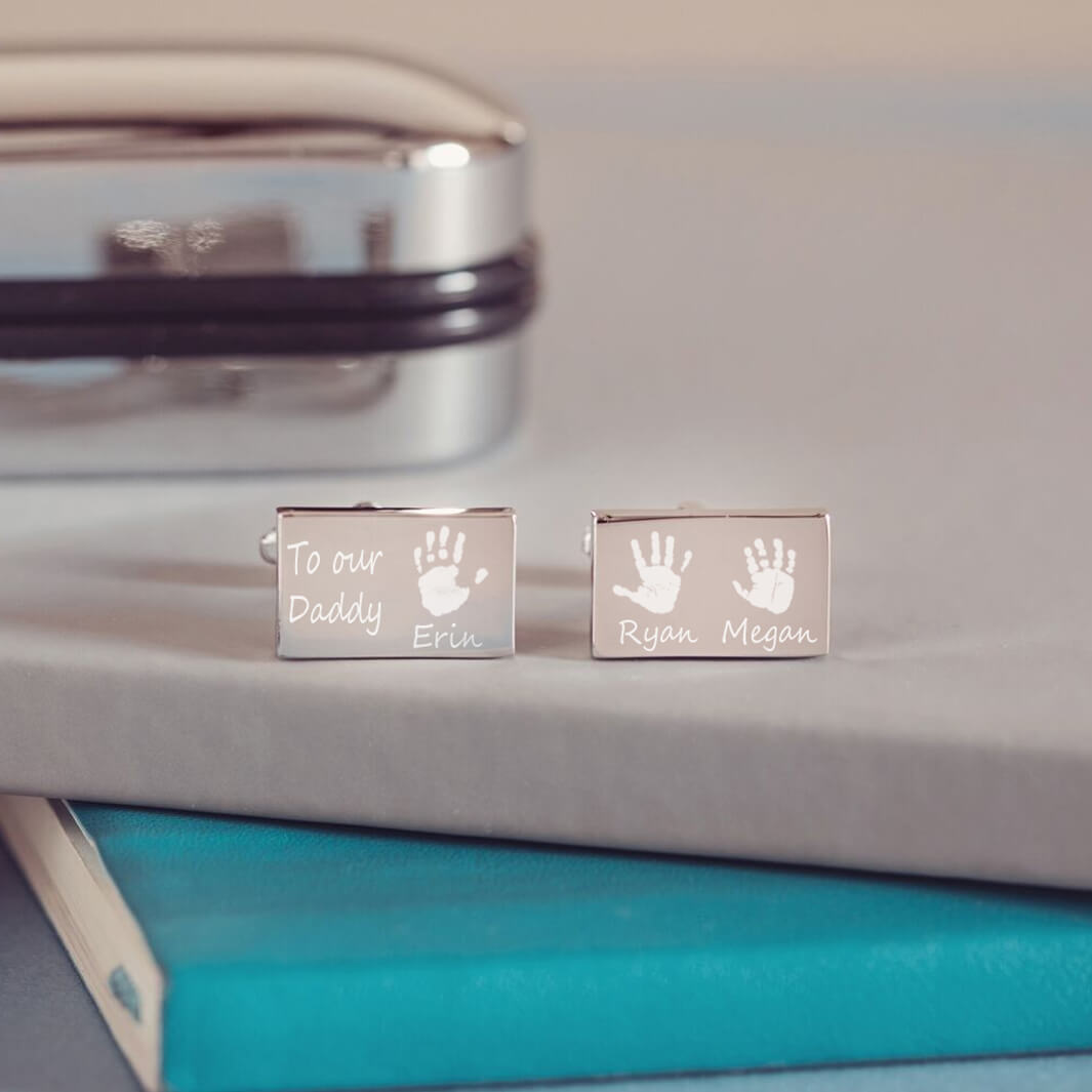 stainless steel rectangular cufflinks with three handprints and names, as well as a short message of 'To Our Daddy' engraved onto the front of the cufflinks. Displayed on a blue and grey background