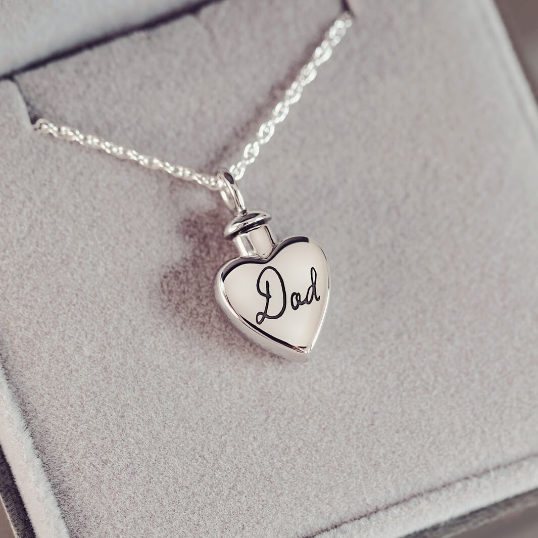 A silver, heart shaped self-fill urn necklace, on a silver chain, with a handwritten message of 'Dad' engraved into the front. Displayed on a grey background