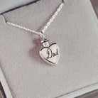 A silver, heart shaped self-fill urn necklace, on a silver chain, with a handwritten message of 'Dad' engraved into the front. Displayed on a grey background